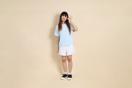 Full body portrait of a young Asian woman wearing a light blue polo shirt, and white shorts, thumbs up, ok or confirm against a plain beige background. College student, Gen Zの写真素材