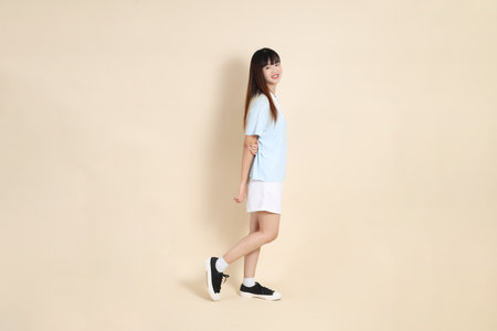 Full body portrait of a young Asian woman wearing a light blue polo shirt, and white shorts, standing with arms crossed and smiling confidently against a plain beige background. College student, Gen Zの写真素材