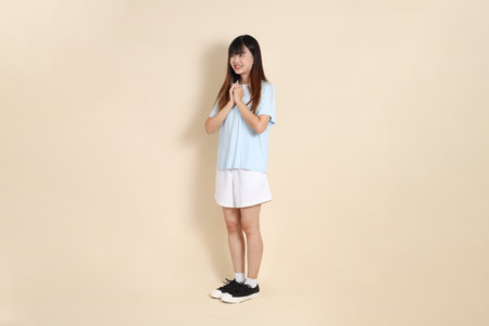 Full body portrait of a young Asian woman wearing a light blue polo shirt, and white shorts, Praying and Peace against a plain beige background. College student, Gen Zの写真素材