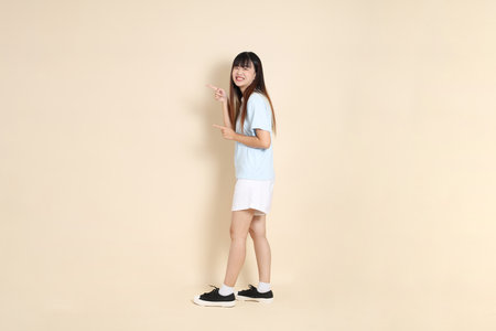 Young Asian woman wearing a light blue polo shirt, and white shorts, pointing against a plain beige background. College student, Gen Z, Summerの写真素材