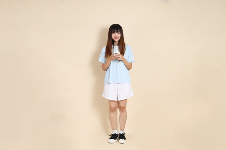 Young Asian woman wearing a light blue polo shirt, and white shorts, Holding mobile phone against a plain beige background. College student, Gen Z, Summerの写真素材