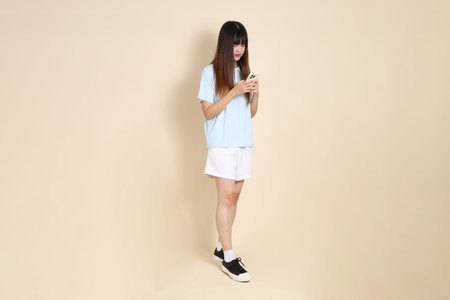 Young Asian woman wearing a light blue polo shirt, and white shorts, Holding mobile phone against a...