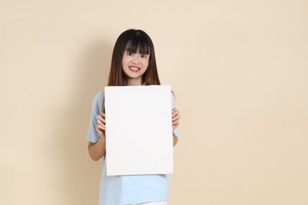 Young Asian woman wearing a light blue polo shirt, and white shorts, Holding blank canvas against a plain beige background. College student, Gen Z, Summerの写真素材