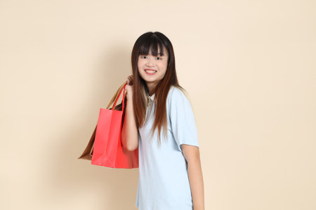 Young Asian woman wearing a light blue polo shirt, holding shopping bag against a plain beige background. College student, Gen Z, Summerの写真素材