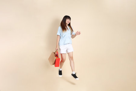 Young Asian woman wearing a light blue polo shirt, and white shorts, holding shopping bag against a plain beige background. College student, Gen Z, Summerの写真素材