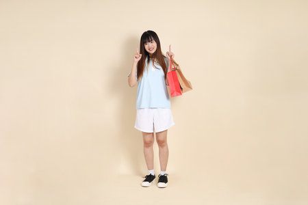 Young Asian woman wearing a light blue polo shirt, and white shorts, holding shopping bag against a plain beige background. College student, Gen Z, Summerの写真素材