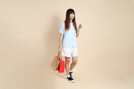 Young Asian woman wearing a light blue polo shirt, and white shorts, holding shopping bag against a plain beige background. College student, Gen Z, Summerの写真素材