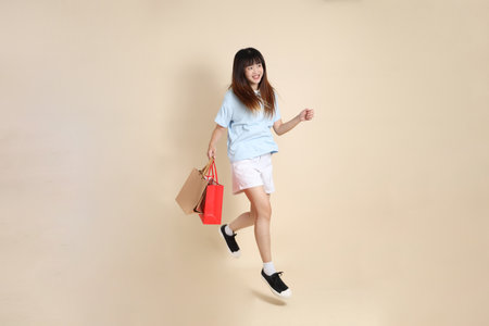 Young Asian woman wearing a light blue polo shirt, and white shorts, holding shopping bag against a plain beige background. College student, Gen Z, Summerの写真素材