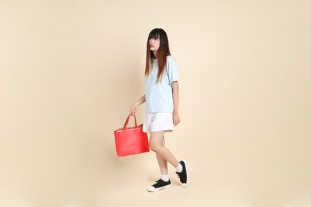 Young Asian woman wearing a light blue polo shirt, and white shorts, holding shopping bag against a plain beige background. College student, Gen Z, Summerの写真素材