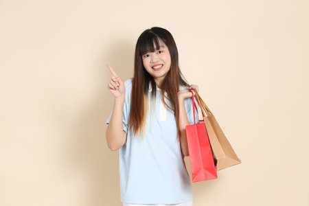 Young Asian woman wearing a light blue polo shirt, and white shorts, holding shopping bag against a plain beige background. College student, Gen Z, Summerの写真素材