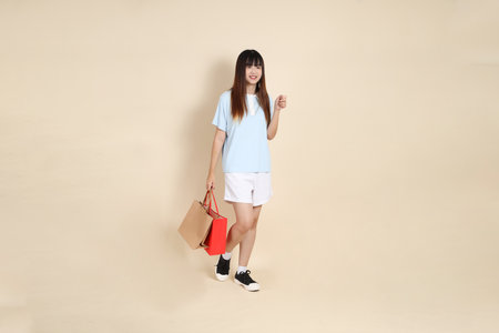 Young Asian woman wearing a light blue polo shirt, and white shorts, holding shopping bag against a plain beige background. College student, Gen Z, Summerの写真素材