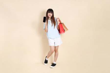 Young Asian woman wearing a light blue polo shirt, and white shorts, holding shopping bag against a plain beige background. College student, Gen Z, Summerの写真素材