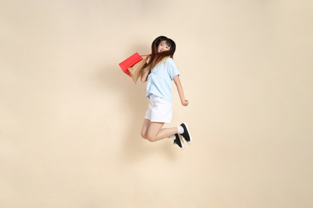 Young Asian woman wearing a light blue polo shirt, and white shorts, holding shopping bag against a plain beige background. College student, Gen Z, Summerの写真素材