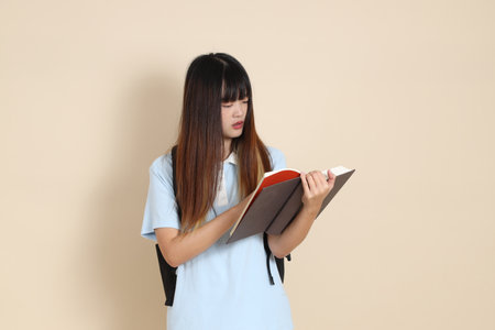 Asian woman wearing blue polo shirt,  carrying school backpack, holding a book against a plain beige background.の写真素材