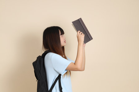 Asian woman wearing blue polo shirt,  white shorts, carrying school backpack, holding a book against a plain beige background. Gen Z,の写真素材