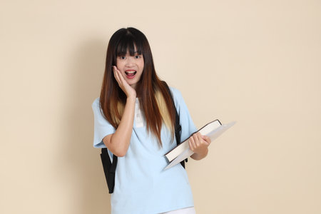 Asian woman wearing blue polo shirt,  white shorts, carrying school backpack, holding a book against a plain beige background. Gen Z,の写真素材