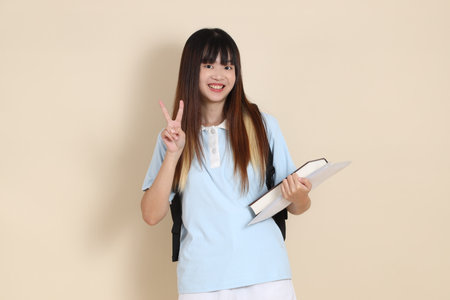 Asian woman wearing blue polo shirt,  white shorts, carrying school backpack, holding a book against a plain beige background. Gen Z,の写真素材