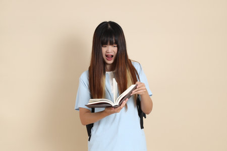 Asian woman wearing blue polo shirt,  white shorts, carrying school backpack, holding a book against a plain beige background. Gen Z,の写真素材