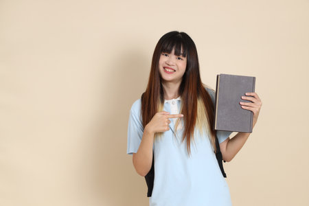 Asian woman wearing blue polo shirt, carrying school backpack, holding a book against a plain beige background. Gen Z,の写真素材