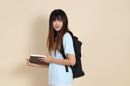 Asian woman wearing blue polo shirt,  white shorts, carrying school backpack, holding a book against a plain beige background. Gen Z,の写真素材