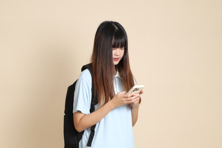 Asian woman wearing blue polo shirt, carrying school backpack, Holding mobile phone against a plain beige background. Gen Z,の写真素材