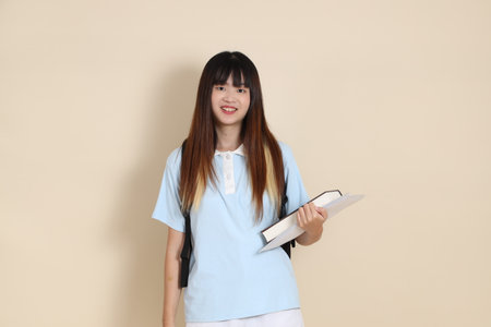 Asian woman wearing blue polo shirt,  white shorts, carrying school backpack, holding a book against a plain beige background. Gen Z,の写真素材