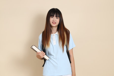 Asian woman wearing blue polo shirt,  white shorts, carrying school backpack, holding a book against a plain beige background. Gen Z,の写真素材