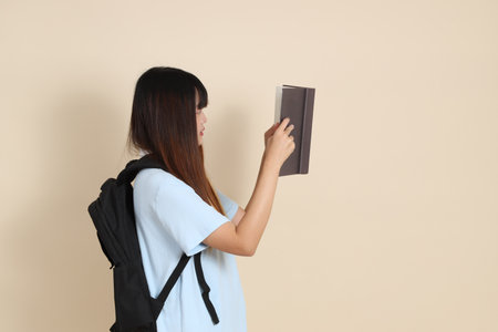 Asian woman wearing blue polo shirt,  white shorts, carrying school backpack, holding a book against a plain beige background. Gen Z,の写真素材