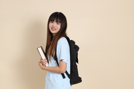 Asian woman wearing blue polo shirt, carrying school backpack, holding a book against a plain beige background. Gen Z,の写真素材
