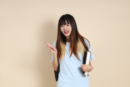 Asian woman wearing blue polo shirt, carrying school backpack, holding a book against a plain beige background. Gen Z,の写真素材