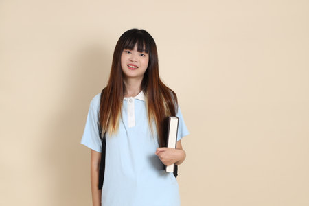 Asian woman wearing blue polo shirt, carrying school backpack, holding a book against a plain beige background. Gen Z,の写真素材
