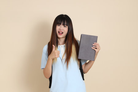 Asian woman wearing blue polo shirt,  white shorts, carrying school backpack, holding a book against a plain beige background. Gen Z,の写真素材