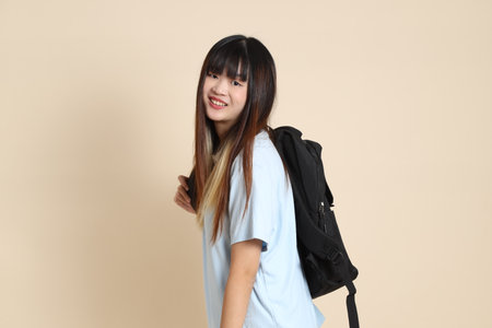 Asian woman wearing blue polo shirt, carrying school backpack, holding a book against a plain beige background. Gen Z,の写真素材