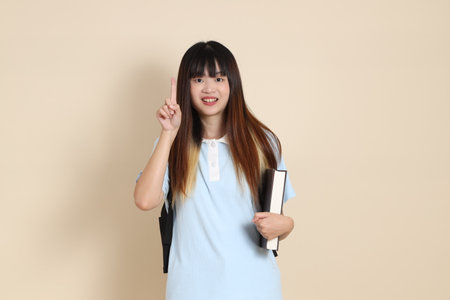Asian woman wearing blue polo shirt,  carrying school backpack, holding a book against a plain beige background. Gen Z,の写真素材