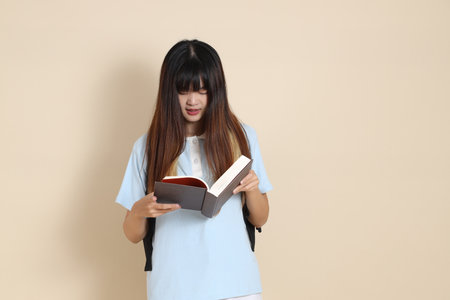 Asian woman wearing blue polo shirt,  white shorts, carrying school backpack, holding a book against a plain beige background. Gen Z,の写真素材