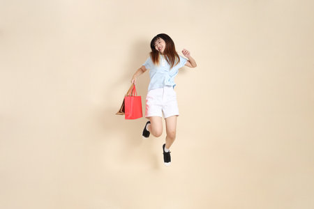 Young Asian woman wearing a light blue polo shirt, and white shorts, holding shopping bag against a plain beige background. College student, Gen Z, Summerの写真素材