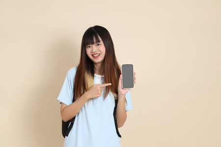 Asian woman wearing blue polo shirt, carrying school backpack, Holding mobile phone against a plain beige background. Gen Z,の写真素材