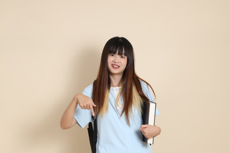 Asian woman wearing blue polo shirt, carrying school backpack, holding a book against a plain beige background. Gen Z,の写真素材