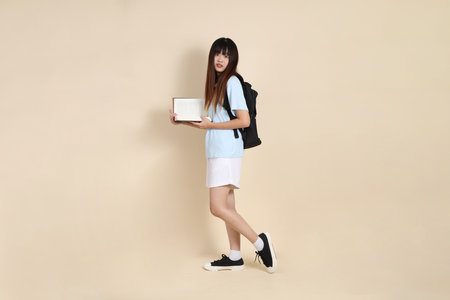 Asian woman wearing blue polo shirt,  white shorts, carrying school backpack, holding a book against a plain beige background. Gen Z,の写真素材