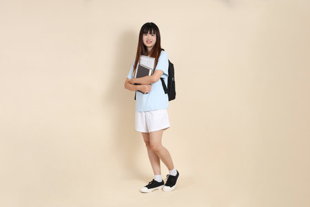 Asian woman wearing blue polo shirt,  white shorts, carrying school backpack, holding a book against a plain beige background. Gen Z,の写真素材