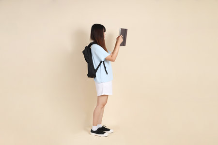 Asian woman wearing blue polo shirt,  white shorts, carrying school backpack, holding a book against a plain beige background. Gen Z,の写真素材