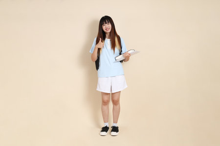 Asian woman wearing blue polo shirt,  white shorts, carrying school backpack, holding a book against a plain beige background. Gen Z,の写真素材