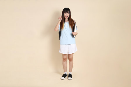 Asian woman wearing blue polo shirt,  white shorts, carrying school backpack, holding a book against a plain beige background. Gen Z,の写真素材