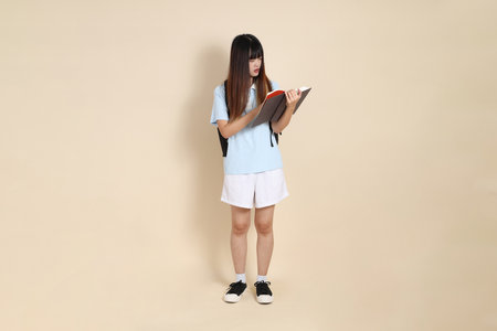 Asian woman wearing blue polo shirt,  white shorts, carrying school backpack, holding a book against a plain beige background. Gen Z,の写真素材