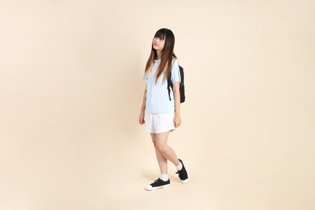 Asian woman wearing blue polo shirt,  white shorts, carrying school backpack, holding a book against a plain beige background. Gen Z,の写真素材