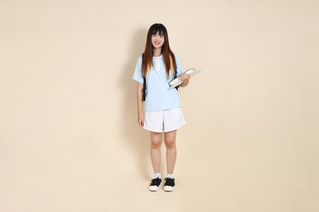 Asian woman wearing blue polo shirt,  white shorts, carrying school backpack, holding a book against a plain beige background. Gen Z,の写真素材