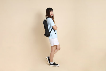 Asian woman wearing blue polo shirt,  white shorts, carrying school backpack, holding a book against a plain beige background. Gen Z,の写真素材