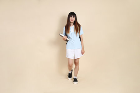 Asian woman wearing blue polo shirt,  white shorts, carrying school backpack, holding a book against a plain beige background. Gen Z,の写真素材