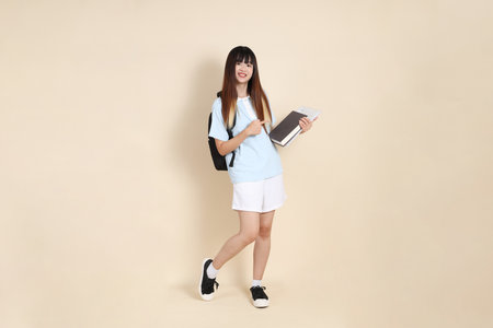 Asian woman wearing blue polo shirt,  white shorts, carrying school backpack, holding a book against a plain beige background. Gen Z,の写真素材