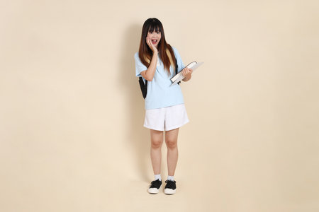 Asian woman wearing blue polo shirt,  white shorts, carrying school backpack, holding a book against a plain beige background. Gen Z,の写真素材